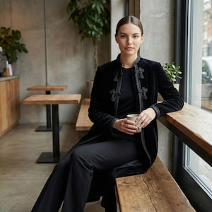 May include: A woman in a black velvet coat with decorative trim and tassels, black trousers, and a black top. She is seated on a wooden bench, holding a cup, in a cafe. The coat is a longline style.