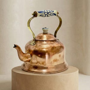 May include: A copper tea kettle with a brass handle and a white and blue floral patterned ceramic handle. The kettle has a rounded body and a spout. The kettle is sitting on a beige surface.