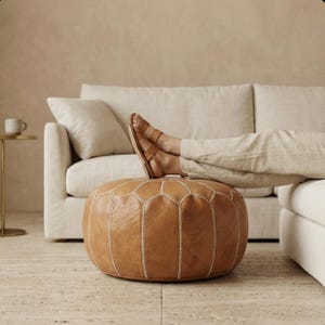 May include: A round, brown leather ottoman with white stitching. The ottoman is in front of a beige sofa. A person is resting their feet on the ottoman, wearing brown sandals and beige pants. The background is a neutral color.