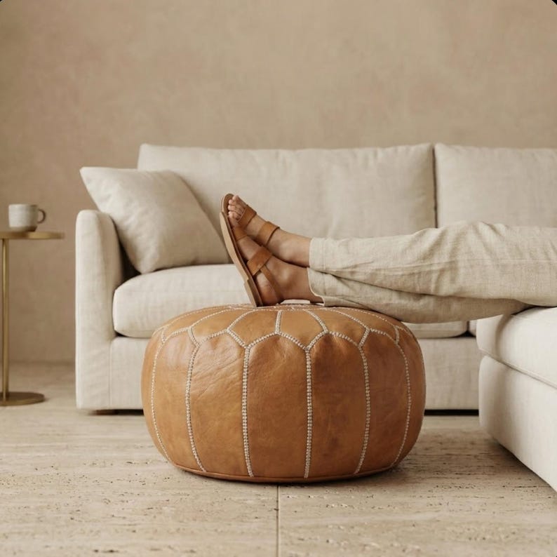 May include: A round, brown leather ottoman with white stitching. The ottoman is in front of a beige sofa. A person is resting their feet on the ottoman, wearing brown sandals and beige pants. The background is a neutral color.