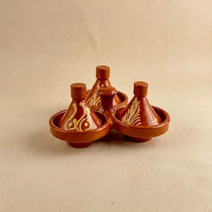 May include: A set of four small, terracotta-coloured tagine pots. Each pot has a conical lid and a round base. The pots are decorated with gold-coloured, hand-painted designs. The tagines are arranged in a circular pattern.