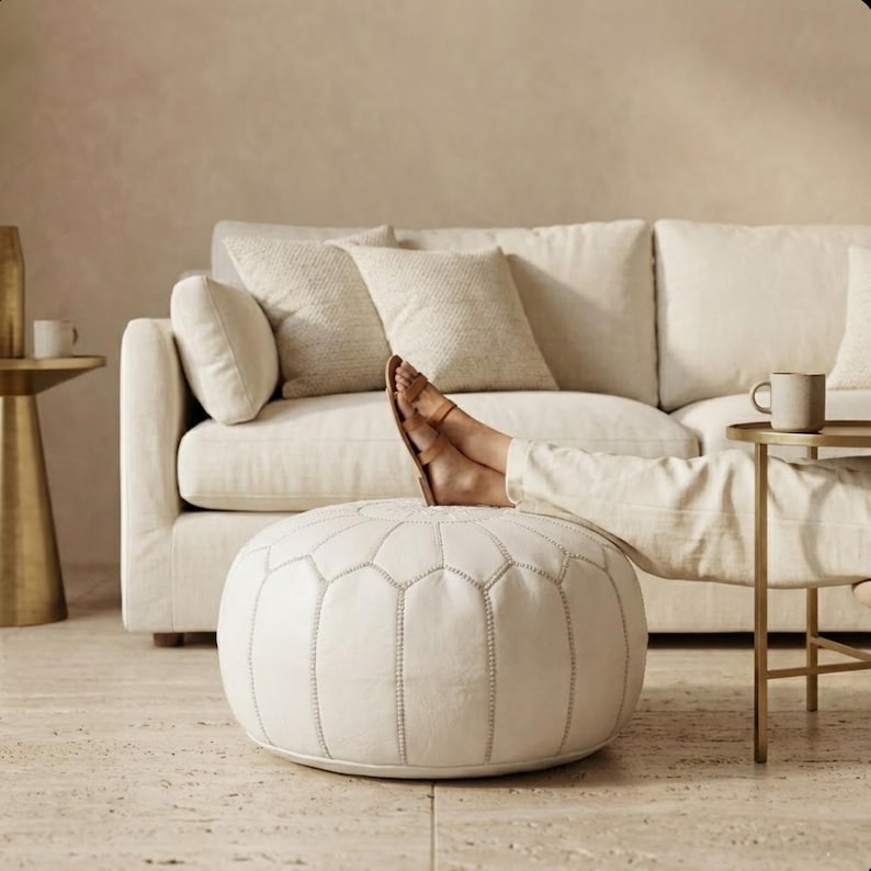 May include: A white, round ottoman with a geometric pattern is the focal point of this image. A person's feet, clad in sandals, rest on the ottoman. A cream-colored sofa and side table with a mug are in the background, creating a relaxing scene.