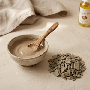 Può includere: Una piccola ciotola di ceramica macchiata piena di una sostanza cremosa marrone chiaro e un cucchiaio di legno. Accanto alla ciotola c'è un mucchio di pezzi di argilla grigi rotti. Una bottiglia di olio di argan è sullo sfondo.
