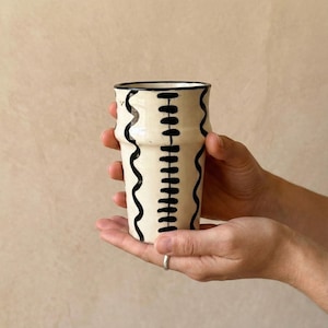 May include: A cream-colored ceramic tumbler with black wavy and vertical line designs. The rim and the top of the tumbler are black. The tumbler is held in both hands against a neutral background.