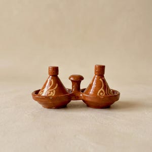 May include: A brown ceramic condiment set featuring two lidded pots with a central handle. Each pot has a conical lid and a decorative swirl design. The set is designed to hold salt and pepper or other spices.