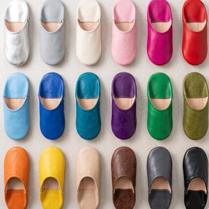 Pode incluir: Uma coleção de chinelos de couro em várias cores, incluindo prata, azul, rosa, vermelho, verde e castanho. Os chinelos estão dispostos em filas, mostrando seus diferentes tons e designs. O calçado parece macio e confortável.