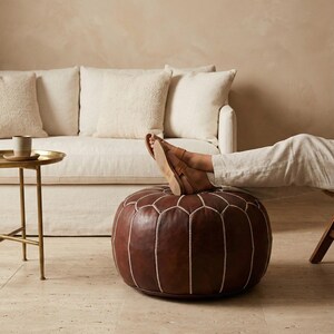 May include: A brown leather ottoman with white stitching, a round shape, and a person's feet resting on top. The ottoman is in front of a white sofa and a small gold side table.