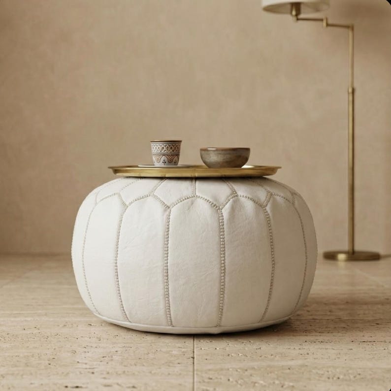 May include: A white, round ottoman with a stitched design. A small gold tray sits on top, holding a decorative cup and a small bowl. The ottoman is in a neutral-toned room with a gold floor lamp.