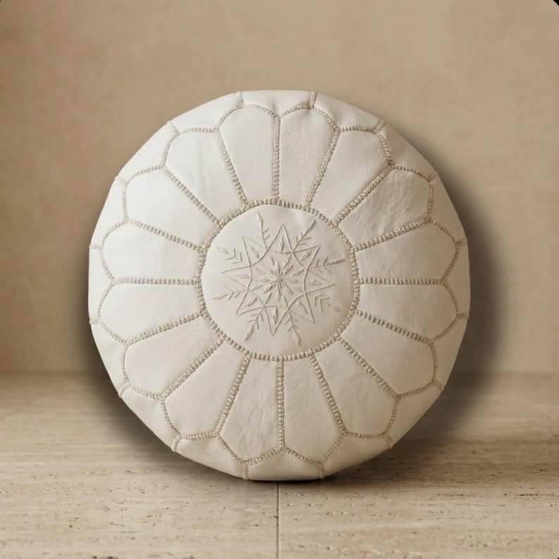 May include: Off-white round Moroccan pouf with a snowflake design embroidered in the center. The pouf is made of leather and has a geometric pattern stitched around the edges. It is a decorative piece for home decor.