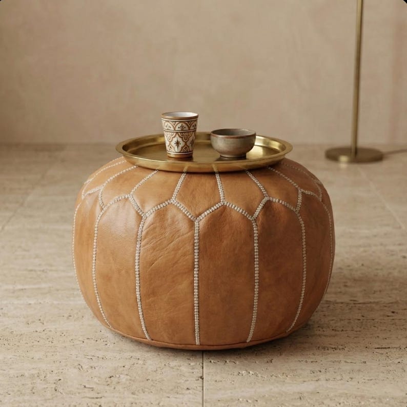 May include: A round, brown leather ottoman with white stitching and a gold-colored tray on top. The tray holds a small patterned cup and a small bowl. The ottoman is a decorative piece of furniture.
