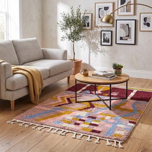 May include: A living room scene featuring a light gray sofa with a mustard-colored throw blanket. A colorful rug with geometric patterns in pink, blue, yellow, and burgundy sits on the wooden floor. A round wooden coffee table and a brass floor lamp complete the decor.