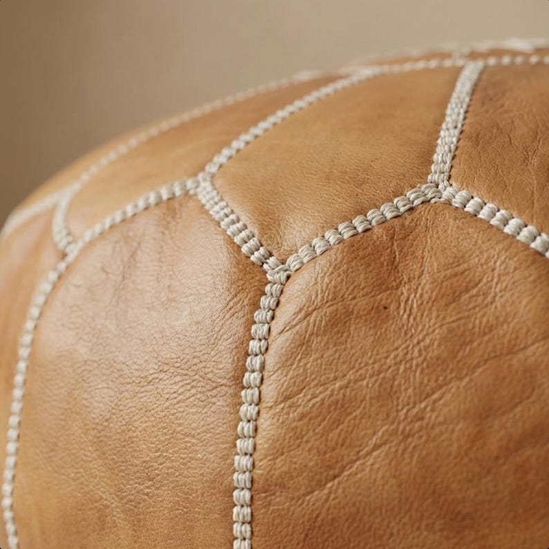 May include: Close-up of a tan leather pouf with a geometric design. The pouf is made of stitched leather panels, with white stitching outlining each panel. The leather has a slightly textured appearance.