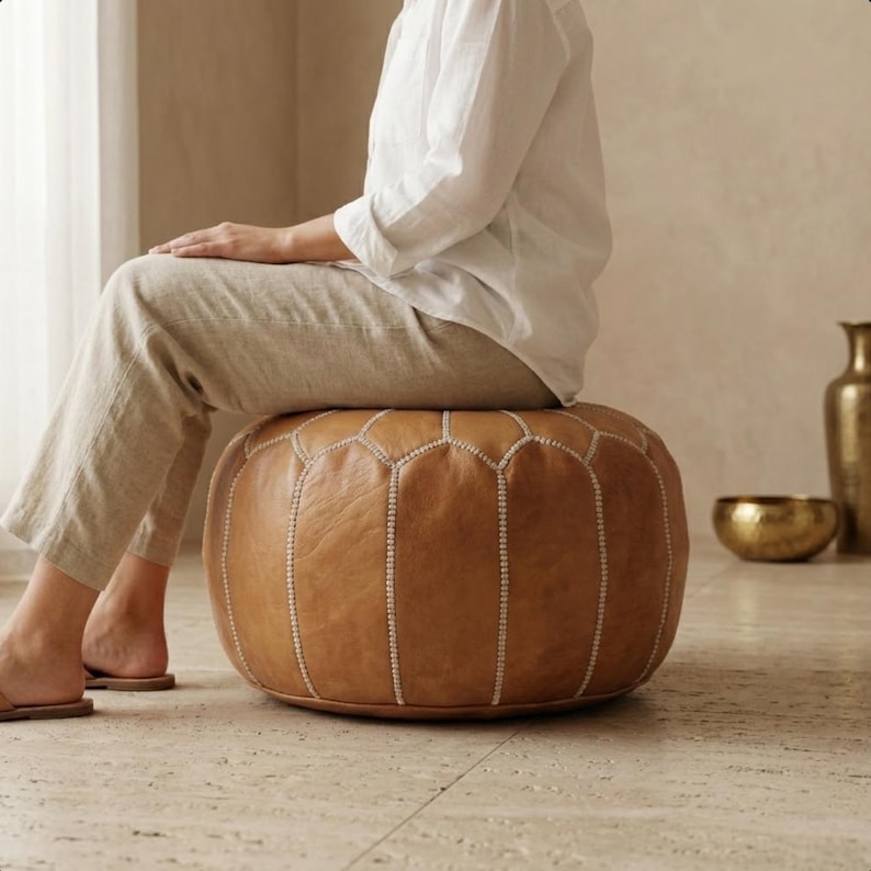 May include: A round, brown leather ottoman with white stitching. The ottoman is a light brown color and has a geometric pattern on the top. The ottoman is a comfortable seating option for a living room or bedroom.