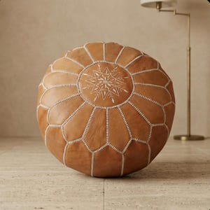 May include: A round, tan leather pouf with a geometric design. The pouf features white stitching and a central embroidered starburst pattern. It is a decorative piece of furniture.