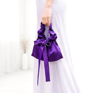 Puede incluir: Un peque&ntilde;o bolso de mano de sat&eacute;n morado con la parte superior fruncida y un gran lazo. El bolso tiene cintas largas y fluidas y lo sostiene una persona que lleva un vestido blanco. El color del bolso es un morado intenso y rico.