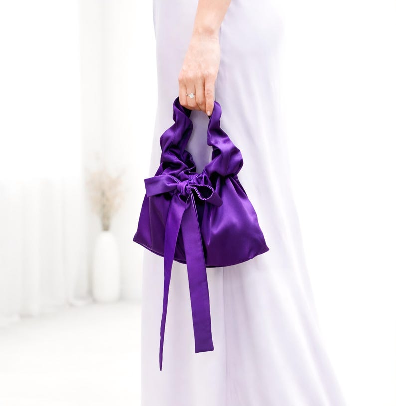 Puede incluir: Un peque&ntilde;o bolso de mano de sat&eacute;n morado con la parte superior fruncida y un gran lazo. El bolso tiene cintas largas y fluidas y lo sostiene una persona que lleva un vestido blanco. El color del bolso es un morado intenso y rico.