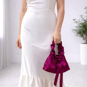 Puede incluir: Un vestido blanco largo, con dobladillo con volantes, se combina con un bolso de mano de sat&eacute;n magenta oscuro. El bolso presenta un dise&ntilde;o fruncido y un lazo grande. El vestido tiene un corpi&ntilde;o sencillo y ajustado y tirantes finos.