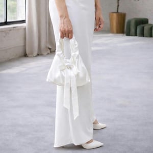 Puede incluir: Un bolso de mano de sat&eacute;n blanco con un lazo y largas cintas es sostenido por una persona que lleva un vestido y zapatos blancos. El bolso tiene forma redondeada y un asa. El fondo es un suelo gris claro y una ventana.