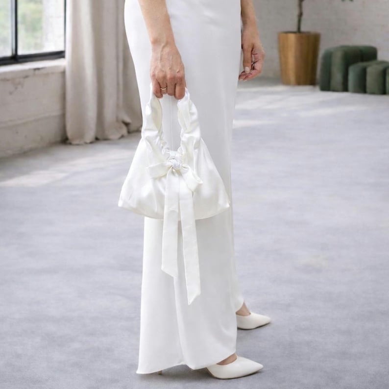 Puede incluir: Un bolso de mano de sat&eacute;n blanco con un lazo y largas cintas es sostenido por una persona que lleva un vestido y zapatos blancos. El bolso tiene forma redondeada y un asa. El fondo es un suelo gris claro y una ventana.