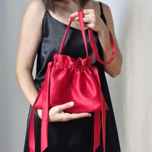 Puede incluir: Un bolso rojo satinado con cordón, asas y lazos de cinta a juego. El bolso tiene una parte superior fruncida y una superficie lisa y reflectante. El bolso lo sostiene una persona que lleva un vestido negro.