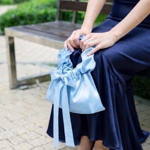 Puede incluir: Un bolso de mano de sat&eacute;n azul claro con cierre de lazo y cintas largas. El bolso lo sostiene una persona que lleva un vestido azul marino y zapatos de tac&oacute;n peep-toe a juego. El fondo incluye un banco de madera y vegetaci&oacute;n.