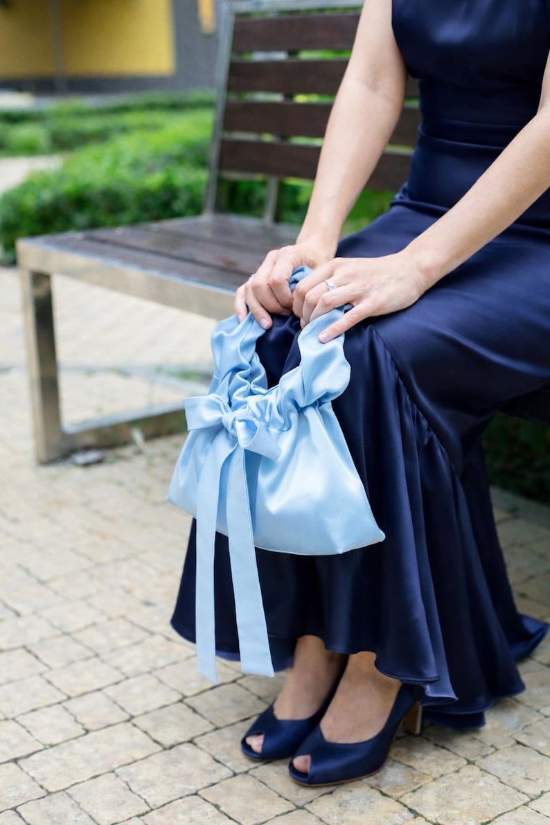 Puede incluir: Un bolso de mano de sat&eacute;n azul claro con cierre de lazo y cintas largas. El bolso lo sostiene una persona que lleva un vestido azul marino y zapatos de tac&oacute;n peep-toe a juego. El fondo incluye un banco de madera y vegetaci&oacute;n.