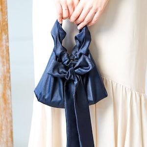 Puede incluir: Un bolso de mano de sat&eacute;n azul marino con un gran lazo y largas cintas. El bolso tiene forma triangular y se sujeta por las asas superiores. Un accesorio elegante.