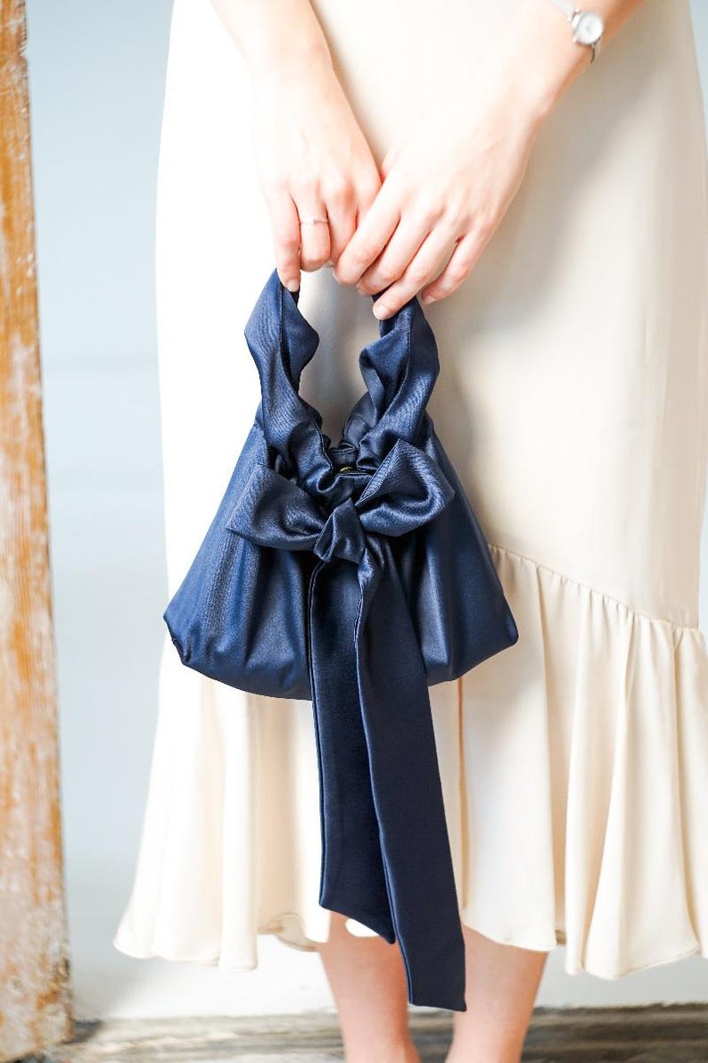Puede incluir: Un bolso de mano de sat&eacute;n azul marino con un gran lazo y largas cintas. El bolso tiene forma triangular y se sujeta por las asas superiores. Un accesorio elegante.