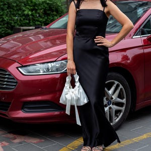 Puede incluir: Un vestido largo negro de sat&eacute;n con tirantes finos. El vestido se combina con un bolso de mano de sat&eacute;n blanco atado con un lazo. Un coche rojo es visible en el fondo.