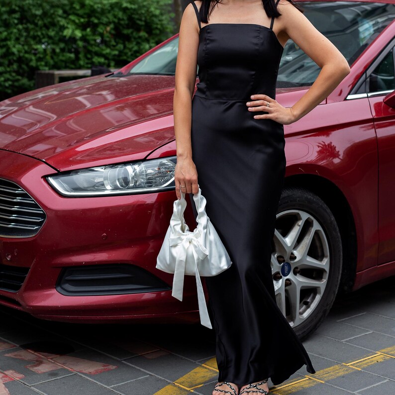 Puede incluir: Un vestido largo negro de sat&eacute;n con tirantes finos. El vestido se combina con un bolso de mano de sat&eacute;n blanco atado con un lazo. Un coche rojo es visible en el fondo.