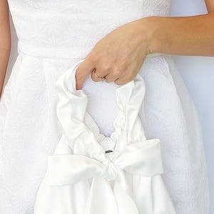 Puede incluir: Un bolso de mano de sat&eacute;n blanco con un lazo grande. El bolso lo sostiene una persona que lleva un vestido blanco. El vestido tiene un estampado floral. El asa del bolso est&aacute; fruncida.