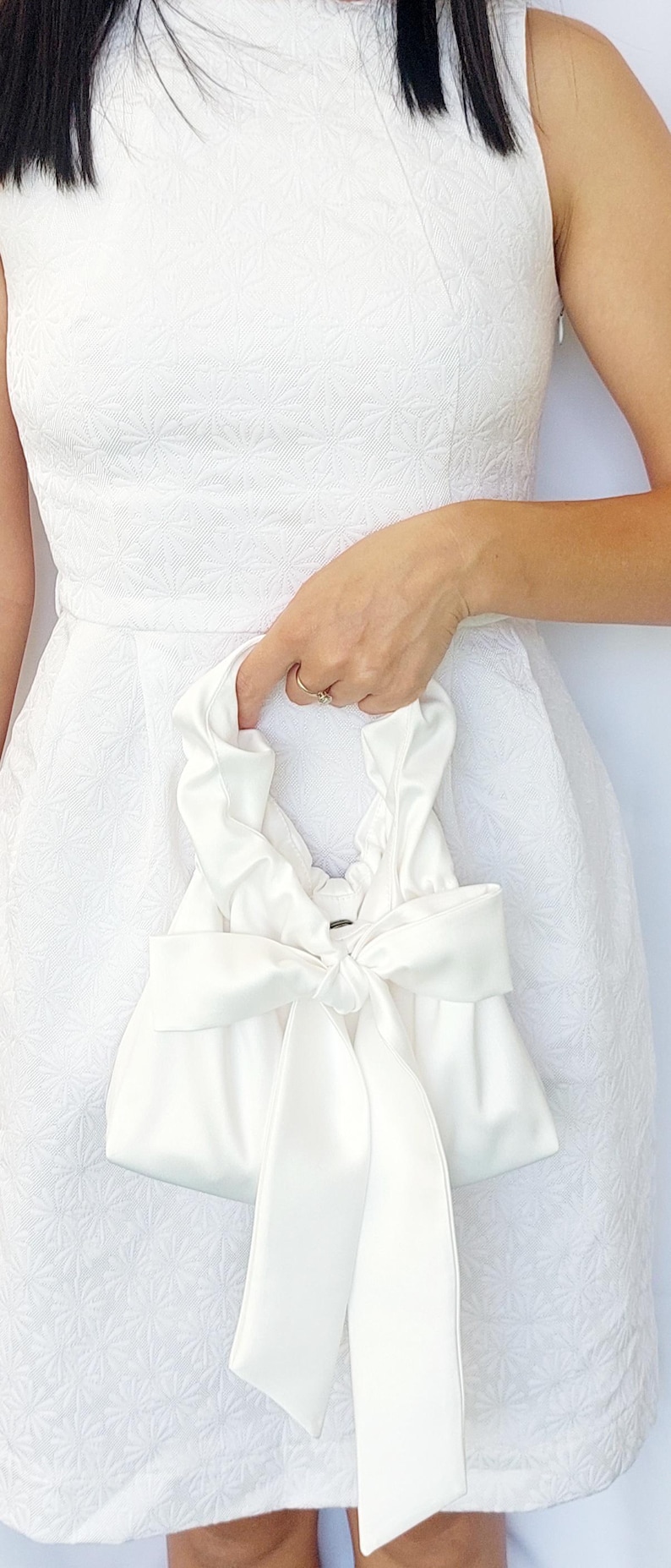 Puede incluir: Un bolso de mano de sat&eacute;n blanco con un lazo grande. El bolso lo sostiene una persona que lleva un vestido blanco. El vestido tiene un estampado floral. El asa del bolso est&aacute; fruncida.