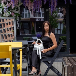 Puede incluir: Una mujer con un vestido negro est&aacute; sentada en una mesa, sosteniendo un bolso de sat&eacute;n blanco con un lazo. La escena es al aire libre, con decoraciones florales y un edificio al fondo. El vestido tiene tirantes finos.