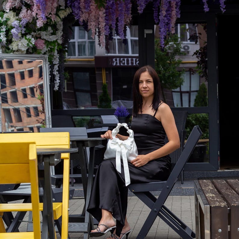 Puede incluir: Una mujer con un vestido negro est&aacute; sentada en una mesa, sosteniendo un bolso de sat&eacute;n blanco con un lazo. La escena es al aire libre, con decoraciones florales y un edificio al fondo. El vestido tiene tirantes finos.