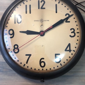 Puede incluir: Reloj de pared General Electric vintage con marco de metal negro. La esfera del reloj es de color crema con números y manecillas negras. El reloj marca las 8:10. El reloj tiene un segundero rojo.