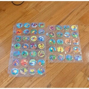May include: Two clear plastic sheets filled with colorful, circular collectible game pieces. Each piece features a unique design, including cartoon characters, sports figures, and text. The pieces are arranged in rows and columns, showcasing a variety of colors and themes.