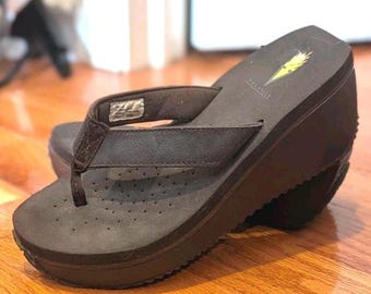 May include: A dark brown wedge flip-flop sandal featuring a thick platform sole, a thong-style strap, and a perforated footbed. The heel area displays a small yellow logo graphic with the text VOLATILE printed in white lettering on the surface.