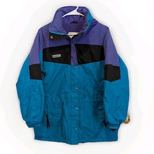 May include: A vintage Columbia jacket in shades of blue, purple, and black. The jacket features a high collar, snap and zipper closure, and a drawstring hem. The Columbia logo is visible on the chest. A stylish and functional outerwear piece.