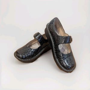 May include: Pair of black Mary Jane style shoes with a glossy, crackled finish. The shoes have a strap with a buckle closure and a small wedge heel. White stitching accents the sole.