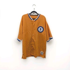 May include: An orange baseball jersey with short sleeves, a button-up front, and a blue and white trim. The jersey features a blue circle patch with a white "N" on the left chest. The sleeves have blue and white trim.