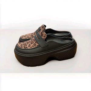 May include: A pair of black platform clogs with a leopard print design on the upper. The clogs have a thick sole and a strap across the top. The brand name is visible on the side of the shoe.