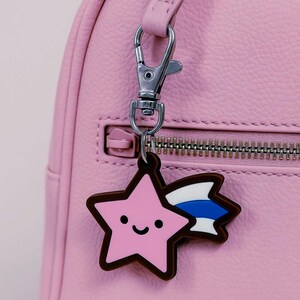 Puede incluir: Un dije en forma de estrella rosa con un contorno marrón y una cara sonriente, sujeto a un cierre plateado. El dije tiene un diseño de cola blanca y azul. El dije cuelga de un bolso rosa.