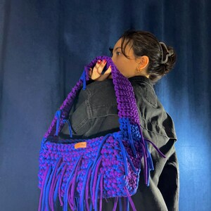 May include: A handmade, large, purple and blue crochet bag with fringe. The bag has a thick strap and is made of a soft, textured material. The fringe is a mix of blue and purple, adding a playful touch to the design.
