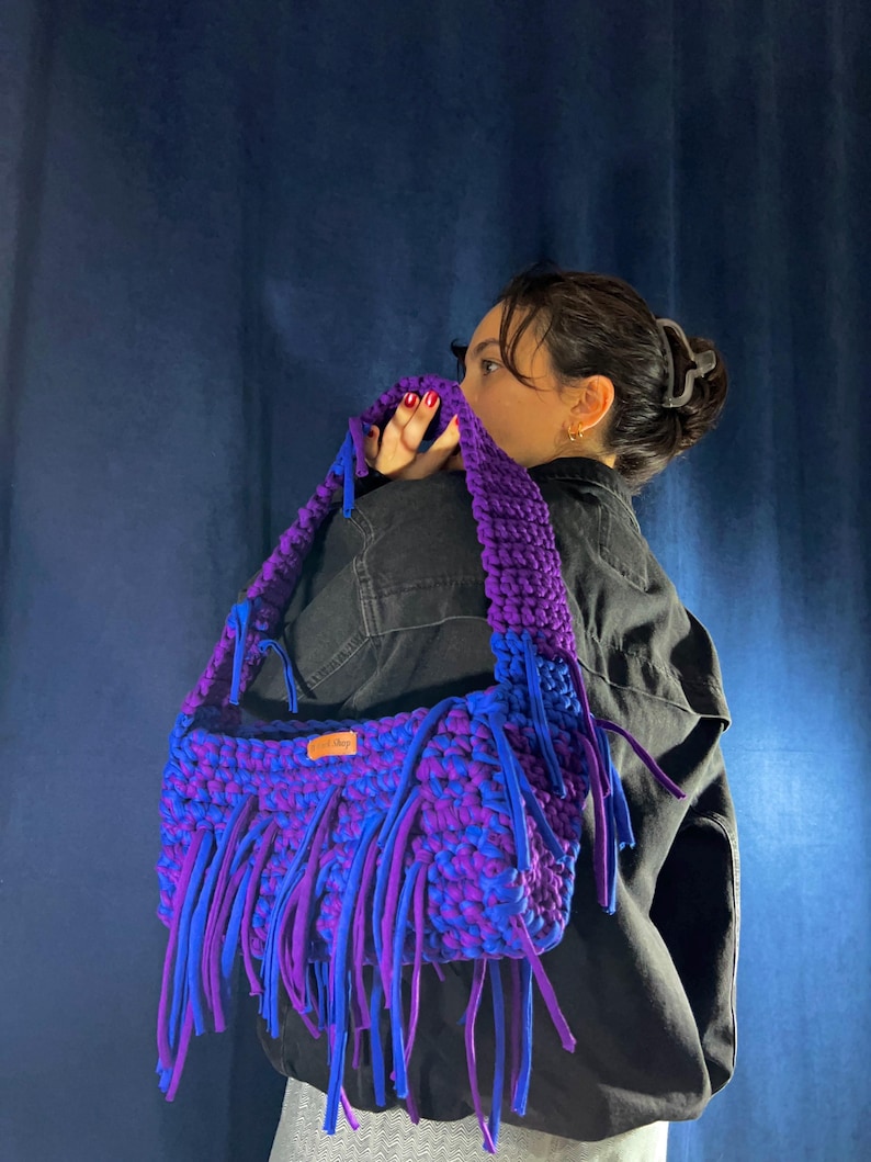 May include: A handmade, large, purple and blue crochet bag with fringe. The bag has a thick strap and is made of a soft, textured material. The fringe is a mix of blue and purple, adding a playful touch to the design.