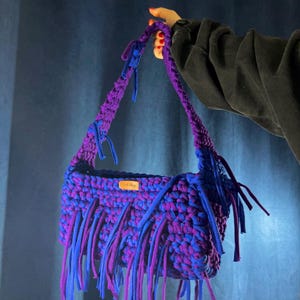 May include: A handmade, crocheted shoulder bag in shades of purple and blue. The bag features a rectangular shape with fringe detailing along the bottom edge. The handles are also crocheted in a matching colour scheme. A small tag is visible on the front.