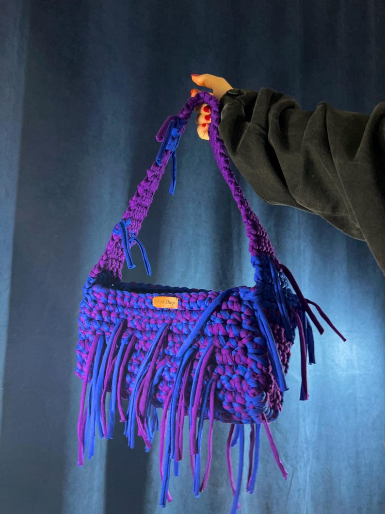 May include: A handmade, crocheted shoulder bag in shades of purple and blue. The bag features a rectangular shape with fringe detailing along the bottom edge. The handles are also crocheted in a matching colour scheme. A small tag is visible on the front.