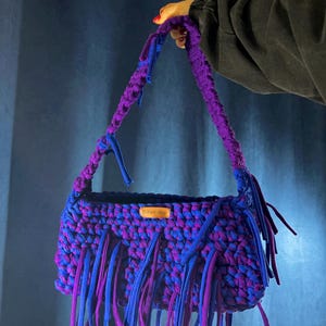 May include: A handmade crochet bag in purple and blue hues. The bag has a thick, braided handle and long fringe detailing. A small wooden tag is attached. The bag is held up by a person.