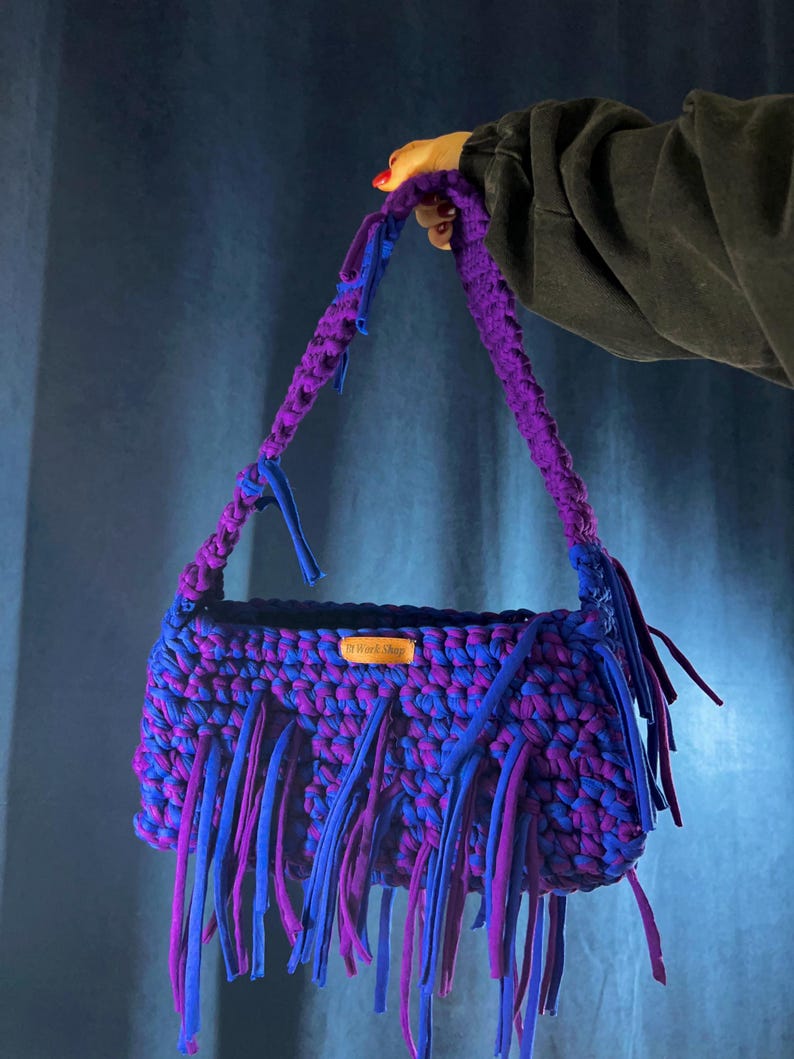 May include: A handmade crochet bag in purple and blue hues. The bag has a thick, braided handle and long fringe detailing. A small wooden tag is attached. The bag is held up by a person.