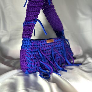 May include: A handmade, crochet handbag in shades of blue and purple. The bag features a rectangular body with a fringed bottom and a wide strap. The colour scheme is a mix of deep purple and bright blue, creating a striking contrast. The bag is made of a soft, textured material.