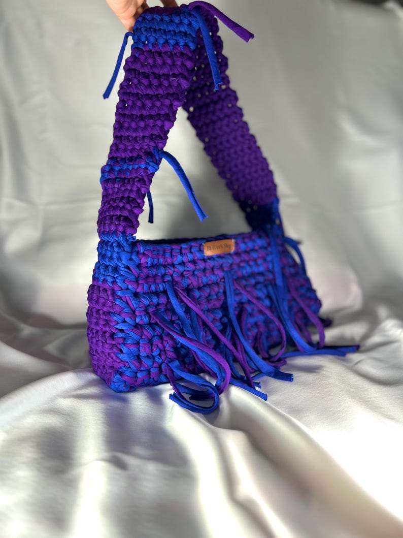 May include: A handmade, crochet handbag in shades of blue and purple. The bag features a rectangular body with a fringed bottom and a wide strap. The colour scheme is a mix of deep purple and bright blue, creating a striking contrast. The bag is made of a soft, textured material.